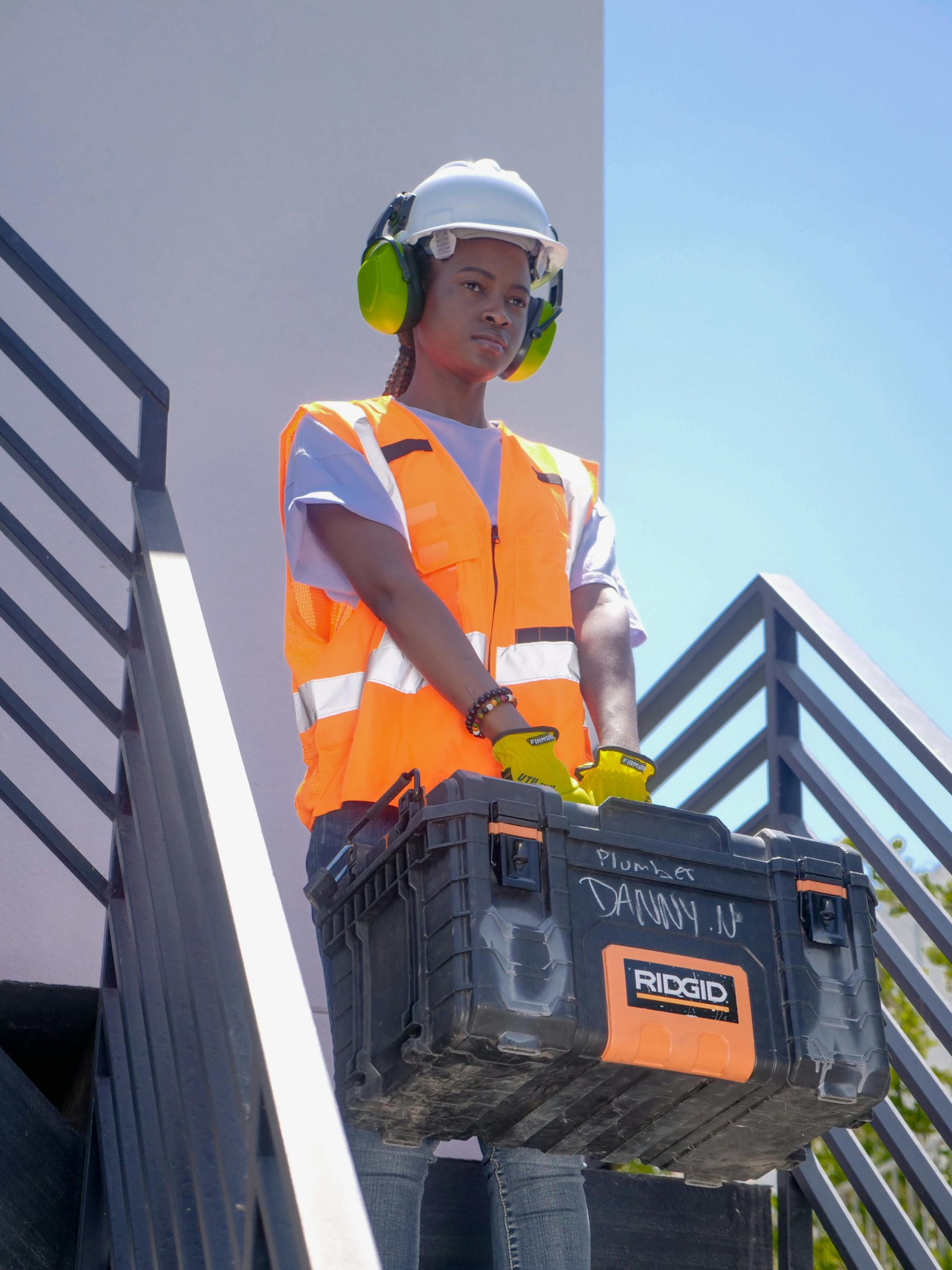 Black female construction worker wearing PPE holds a toolbox on stairs outdoors.